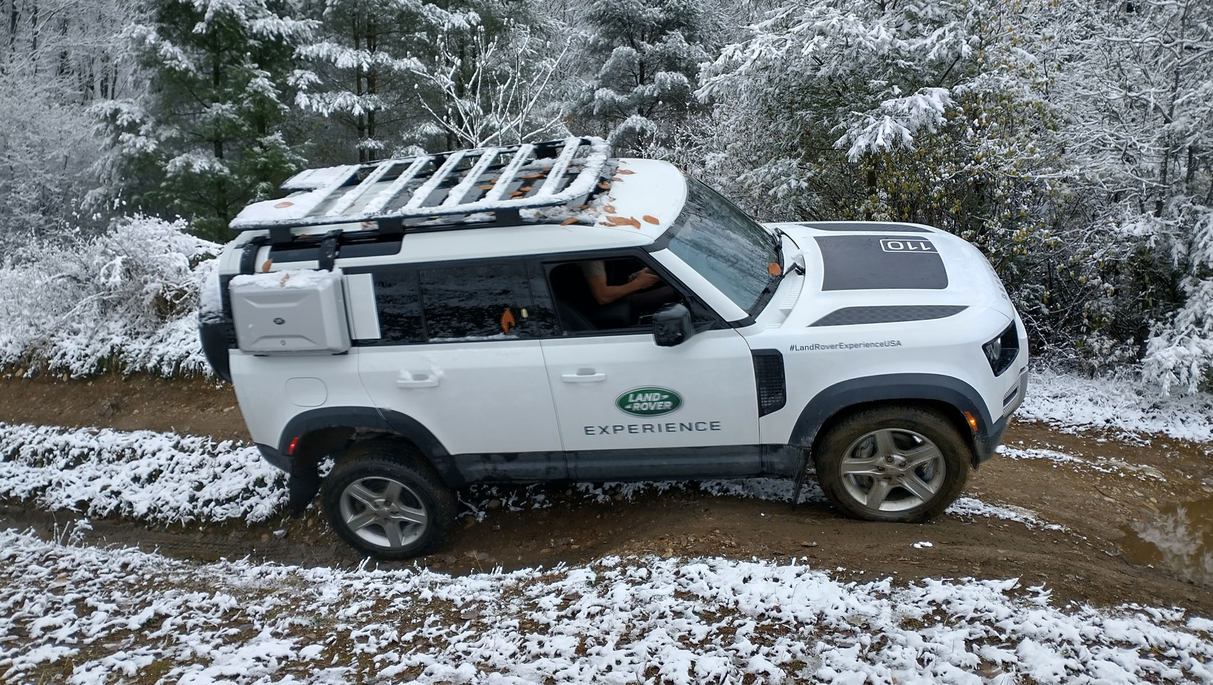 White Land Rover Defender 110 driving on a snowy, muddy off-road trail in a winter forest