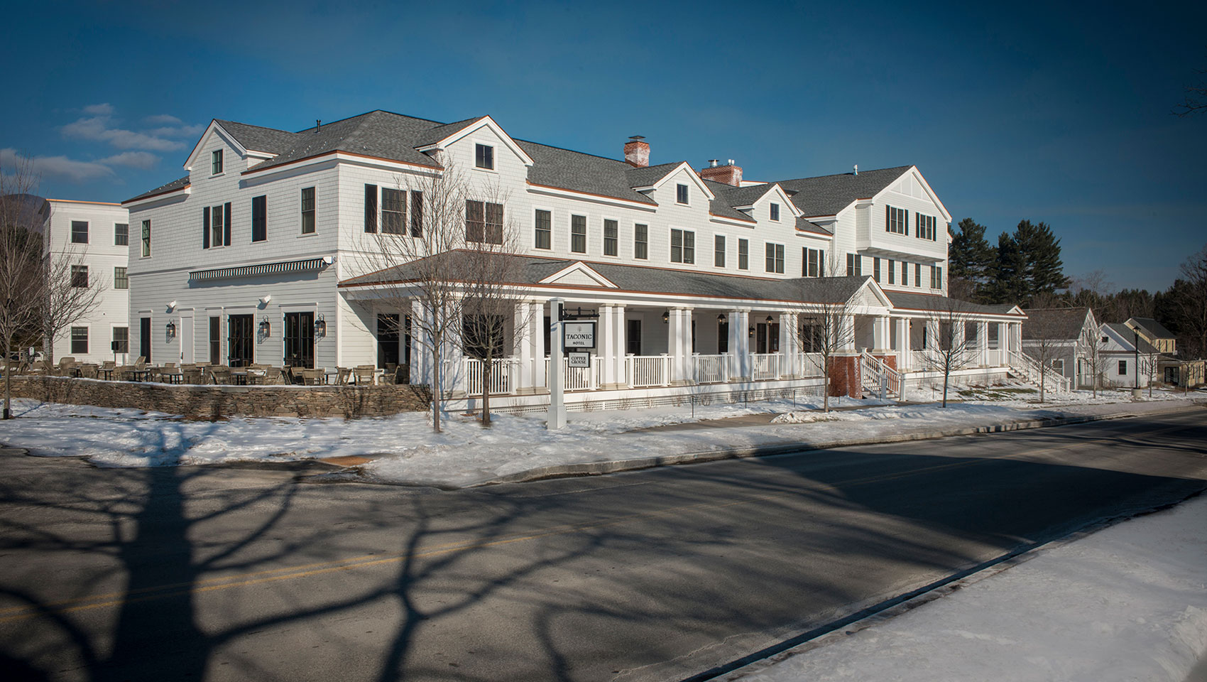 A grand white Taconic Hotel with a long porch and patio amidst snow, shadows, and a street