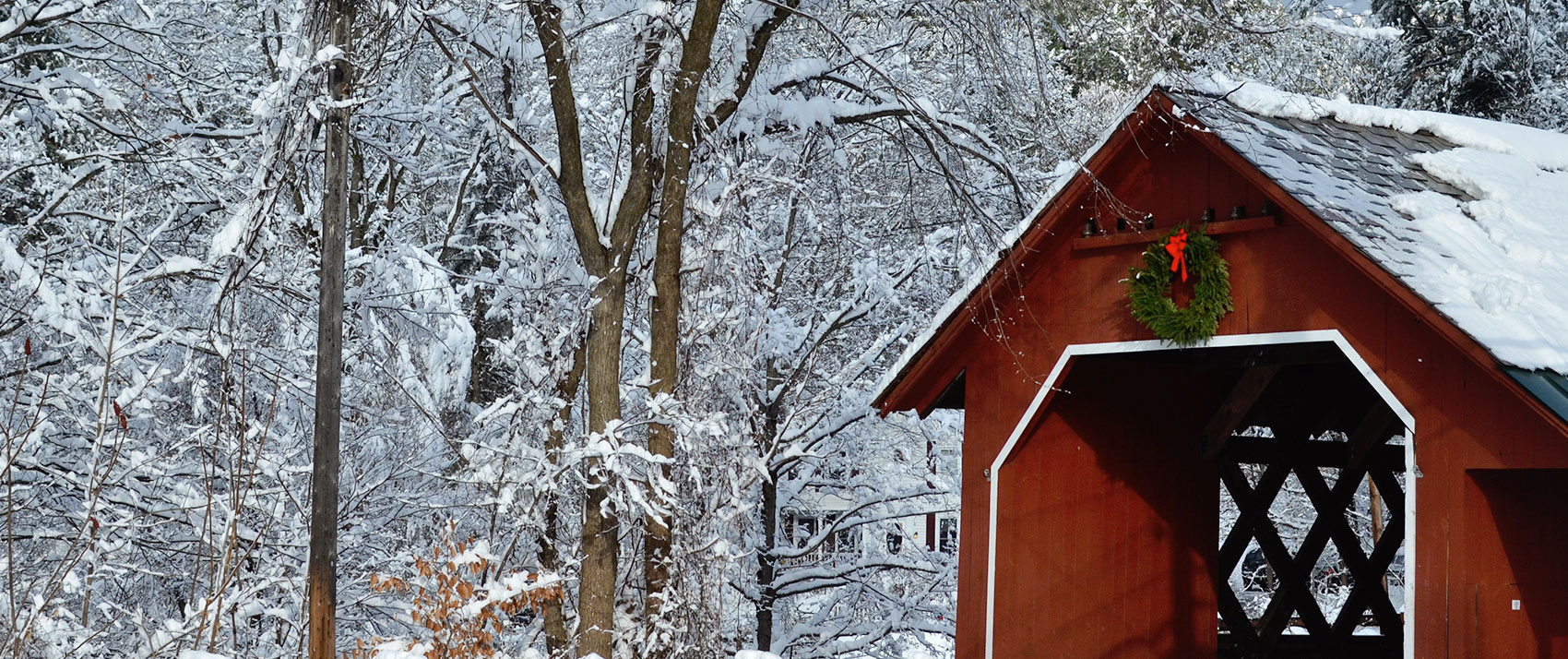 Snow-draped forest framing a red covered bridge with a festive green wreath and red bow